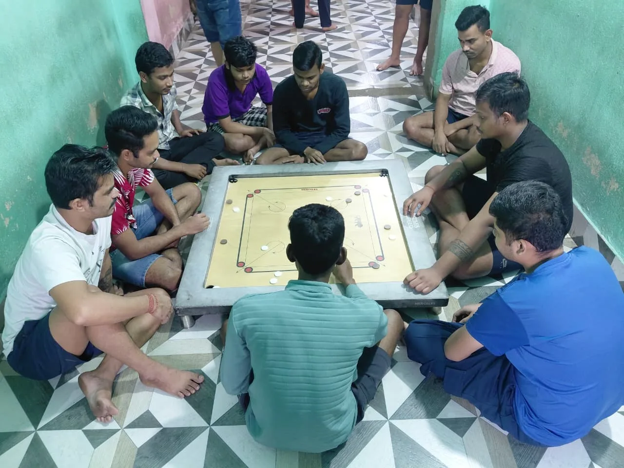Group of men sitting on the floor around a carrom board in a narrow room