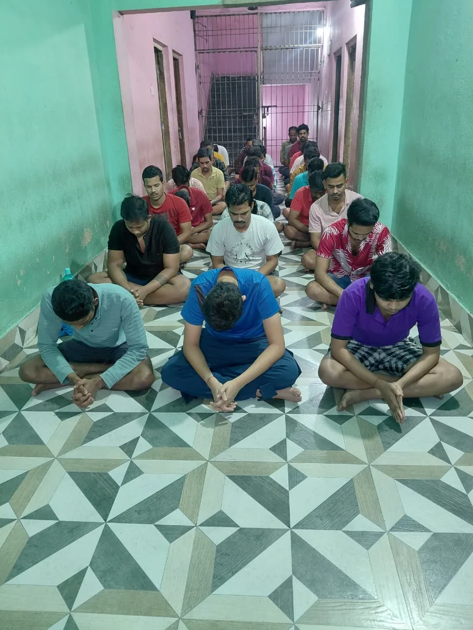 Men sitting cross-legged with bowed heads and clasped hands in a tiled hallway
