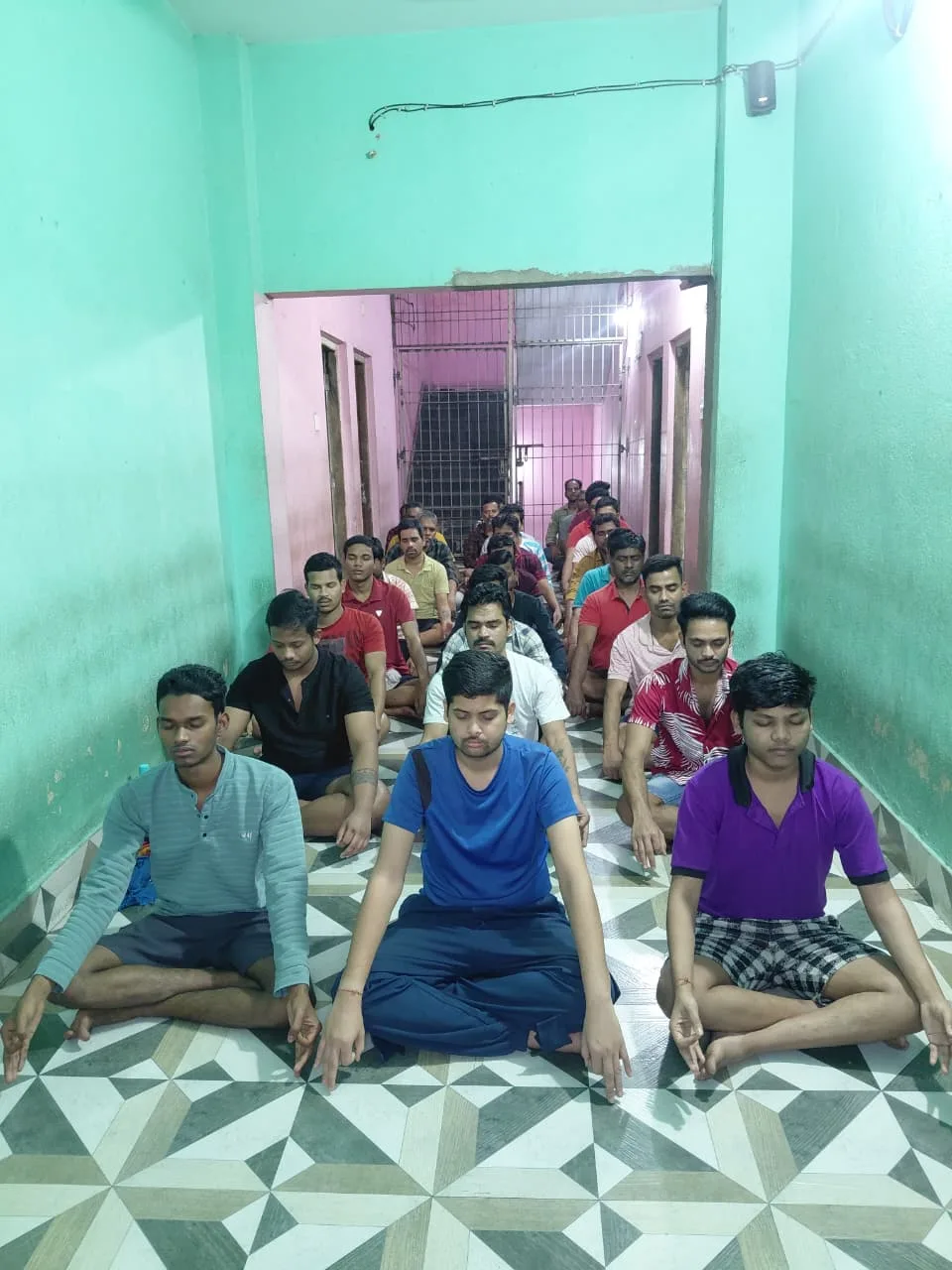 Men meditating cross-legged on a tiled hallway floor inside a mint-green