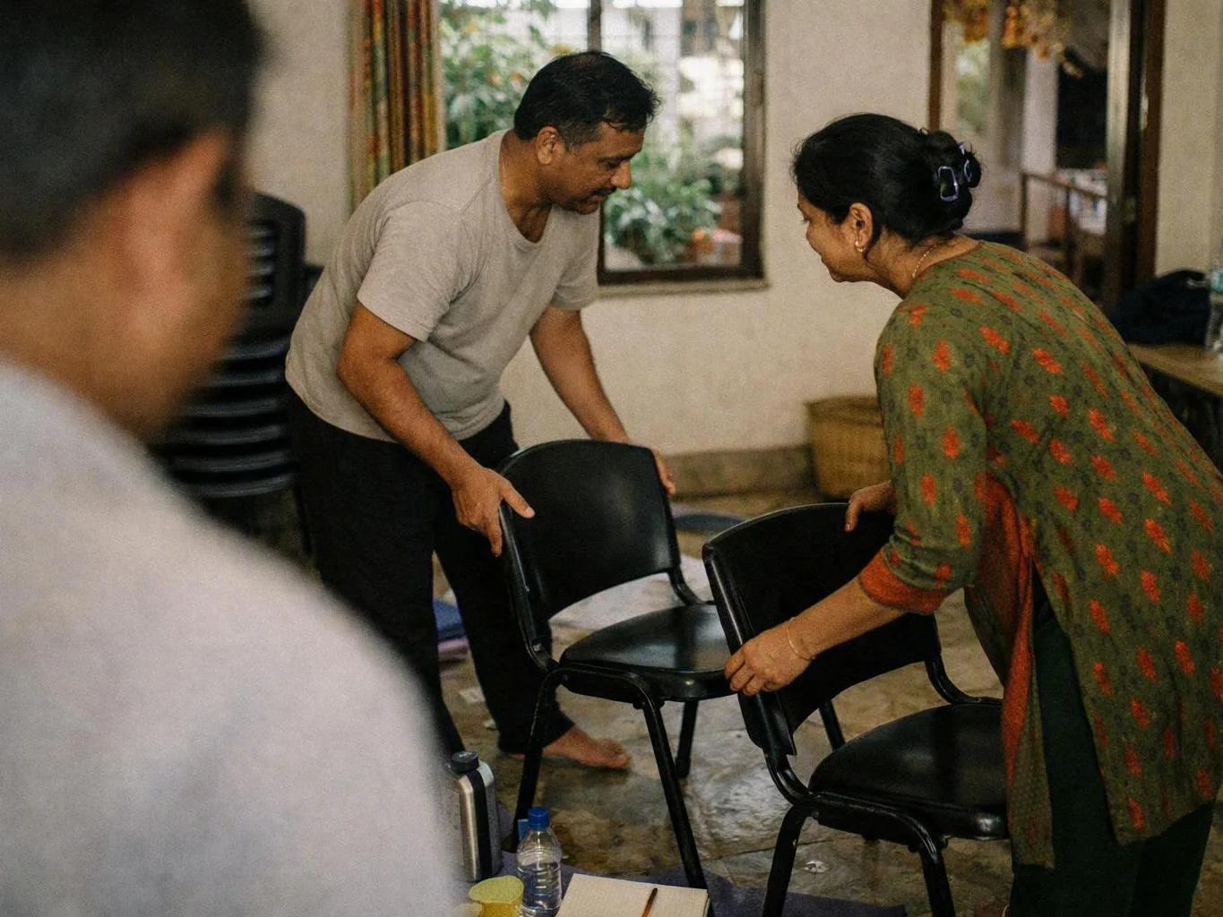 Two adults arrange black chairs in a modest indoor room with windows